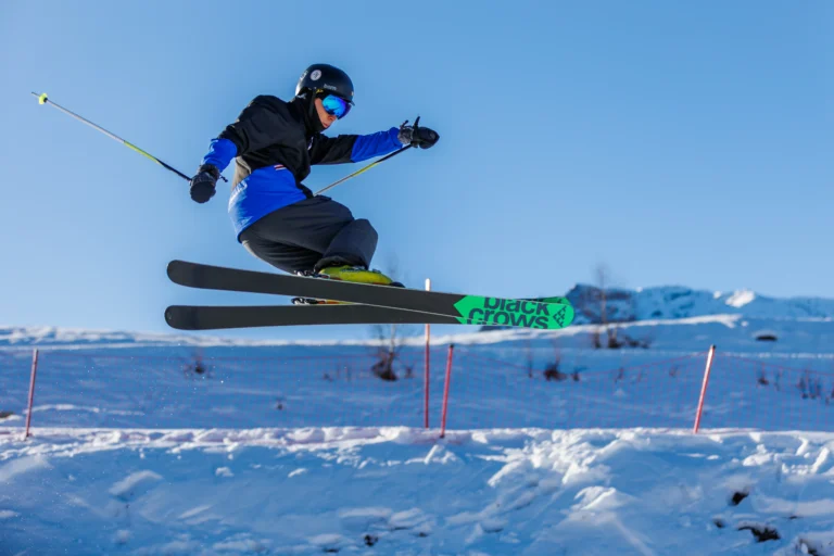 Sciatore in salto nello Snowpark con gli sci incrociati sotto il cielo azzurro.